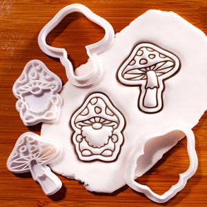 May include: A set of white plastic biscuit cutters featuring two gnome shapes and two mushroom shapes, with two of the cutters pressed into a piece of white dough to show the imprinted outlines of a gnome and a mushroom with detailed caps.
