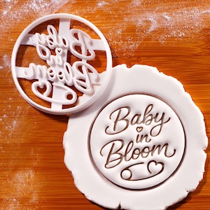 Peut inclure: Emporte-pièce en plastique blanc avec les mots "Baby in Bloom" et un motif de cœur et d'épingle à nourrice. Le découpoir est à côté d'un cercle de pâte blanc avec le même motif imprimé. Le fond est une surface en bois.