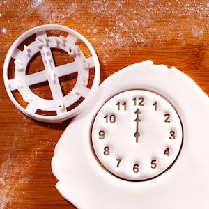 May include: A white plastic clock-shaped cookie cutter next to a white fondant clock. The fondant clock shows the time as 12:00. The cookie cutter is round with the numbers and hands of the clock cut out.