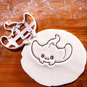May include: A white plastic cookie cutter in the shape of a cartoon manta ray, next to a piece of white dough with the manta ray design pressed into it. The background is a wooden surface.