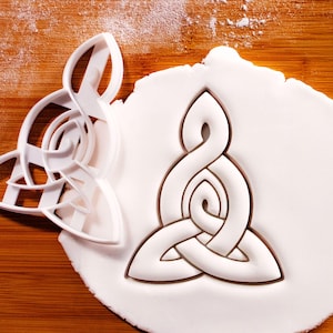 May include: A white plastic cookie cutter shaped like a Celtic knot, alongside a white dough cutout of the same design. The cutter is on a wooden surface, with a dusting of white powder.