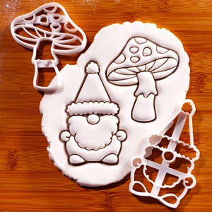 May include: Two white plastic cookie cutters shaped like a gnome and a mushroom, alongside a piece of white rolled dough featuring the pressed outlines of the same gnome and mushroom designs, showcasing the detailed impressions left by the cutters.