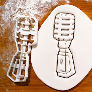 May include: White plastic cookie cutter shaped like a vintage microphone. The cutter is on a white surface with a dusting of flour.