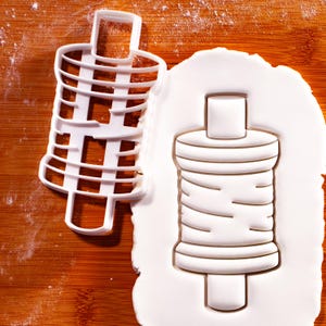 May include: A white plastic cookie cutter in the shape of a thread spool next to a spool-shaped imprint in white dough. The cutter has a grid-like design, and the dough imprint shows the spool's details.