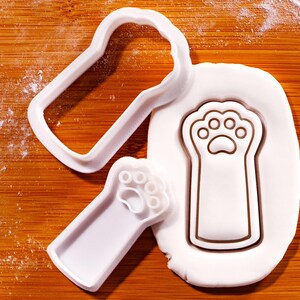 May include: White plastic paw print cookie cutters and a paw print impression in white dough on a wooden surface. The cutters are in the shape of a paw print and a rectangular outline.