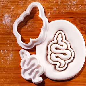 Könnte beinhalten: Weiße Kunststoff-Ausstecher in Schlangen- und Wolkenform sowie weißer Fondant mit Schlangenabdruck. Die Ausstecher und der Fondant liegen auf einer Holzoberfläche, möglicherweise einem Schneidebrett.