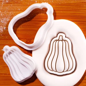 May include: White plastic pumpkin-shaped cookie cutters and a pumpkin-shaped design on a white surface. The cutters are in different sizes and shapes, ideal for autumn baking and crafts.