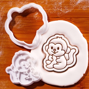 Könnte beinhalten: Weiße, affenförmige Ausstecher und ein affenförmiger Keks auf einer weißen Oberfläche. Der Affen-Ausstecher hat die Form einer Wolke. Der Affen-Keks ist braun und weiß.