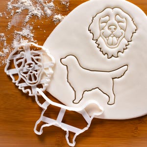 May include: Three white plastic cookie cutters shaped like a golden retriever dog. The cutters are on a wooden surface with white flour scattered around them. The cutters are in different sizes and show the dog's head, full body, and a side profile.