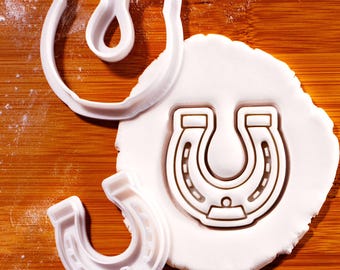 May include: White plastic horseshoe-shaped cookie cutters on a wooden surface. One cutter is pressed into white dough, showing the horseshoe imprint. Two other cutters are next to the dough. The image is well-lit, highlighting the details of the cutters and the dough.