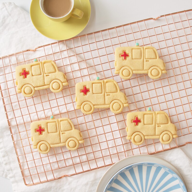 May include: Five yellow ambulance cookie cutters on a copper wire cooling rack. The cookie cutters are shaped like ambulances with red crosses and a light on top.