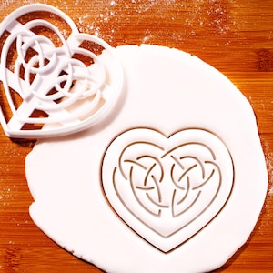 May include: A white plastic heart-shaped cookie cutter with a Celtic knot design, beside a heart-shaped cutout in white dough. The cutter is on a wooden surface with a dusting of white powder.