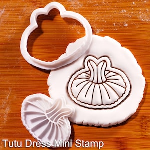 Peut inclure: Un emporte-pièce en plastique blanc et un tampon assorti en forme de robe tutu. Le tampon est pressé dans un morceau de pâte blanche circulaire, créant une impression détaillée du vêtement. Le texte Tutu Dress Mini Stamp est visible.