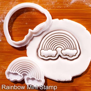 Può includere: Mini timbro arcobaleno in plastica bianca con un taglierino abbinato, accanto a un'impronta arcobaleno su argilla bianca. Il testo "Rainbow Mini Stamp" è in basso nell'immagine. Il timbro presenta un design arcobaleno con nuvole.