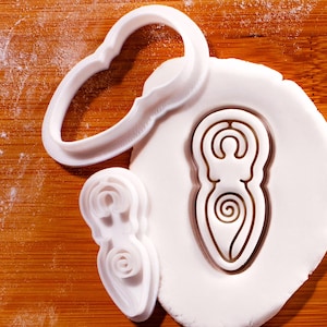 May include: White plastic cookie cutters in the shape of a goddess symbol, a heart, and a smaller goddess symbol are on a wooden surface. The goddess symbol is pressed into white dough. The heart-shaped cutter is at the top.