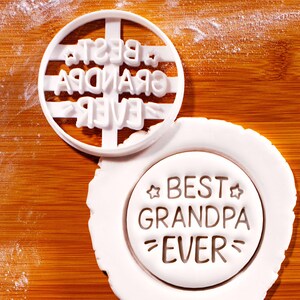 Peut inclure: Emporte-pièce en plastique blanc avec l'inscription "BEST GRANDPA EVER" dans un motif circulaire. Le découpoir est placé sur un morceau de pâte blanche avec le même texte imprimé. Le fond est une surface en bois.