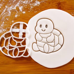 Może przedstawiać: Biała plastikowa wykrawaczka w kształcie żółwia obok dekoracji z masy cukrowej w kształcie żółwia na drewnianej powierzchni. Żółw z masy cukrowej ma uśmiechniętą twarz i segmentowy wzór skorupy. Mąka jest lekko posypana na drewnie.