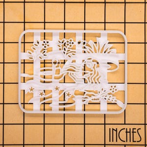 May include: A white plastic cookie cutter with a floral and flowing design. The cutter is rectangular with a grid pattern and the word "INCHES" in the bottom right corner. The design includes flowers, leaves, and a woman's face.