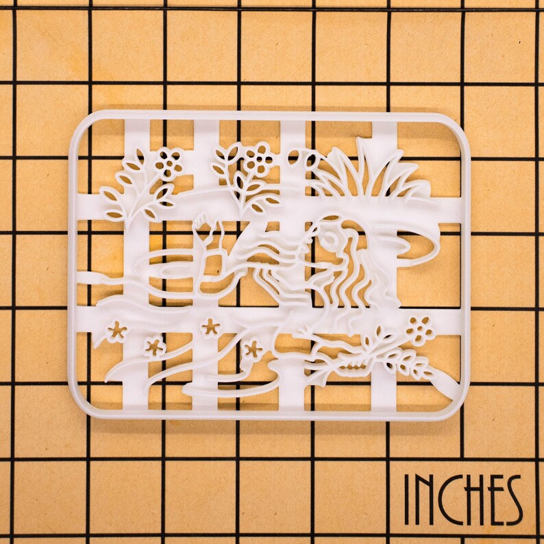 May include: A white plastic cookie cutter with a floral and flowing design. The cutter is rectangular with a grid pattern and the word "INCHES" in the bottom right corner. The design includes flowers, leaves, and a woman's face.