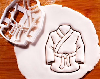 May include: A white plastic cookie cutter shaped like a martial arts uniform with a wrap-around jacket and a tied belt. The cutter is shown next to a piece of rolled-out white dough that has been pressed to reveal the uniform's outline and details.