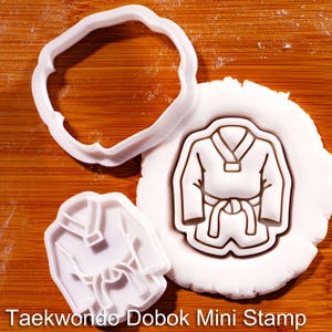 Op de afbeelding: Een witte plastic koekjesvorm en een bijpassende stempel met het ontwerp van een Taekwondo dobok-uniform. De stempel is in een rond stuk wit deeg gedrukt, waardoor een duidelijke omtrek van het vechtsportuniform met een geknoopte riem ontstaat.