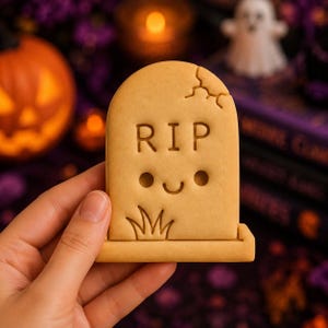 May include: A light brown, tombstone-shaped biscuit featuring the embossed letters RIP, a small smiling face, and a patch of grass at the base. The top of the biscuit has a decorative crack detail, and the surface is smooth and uniform in colour.