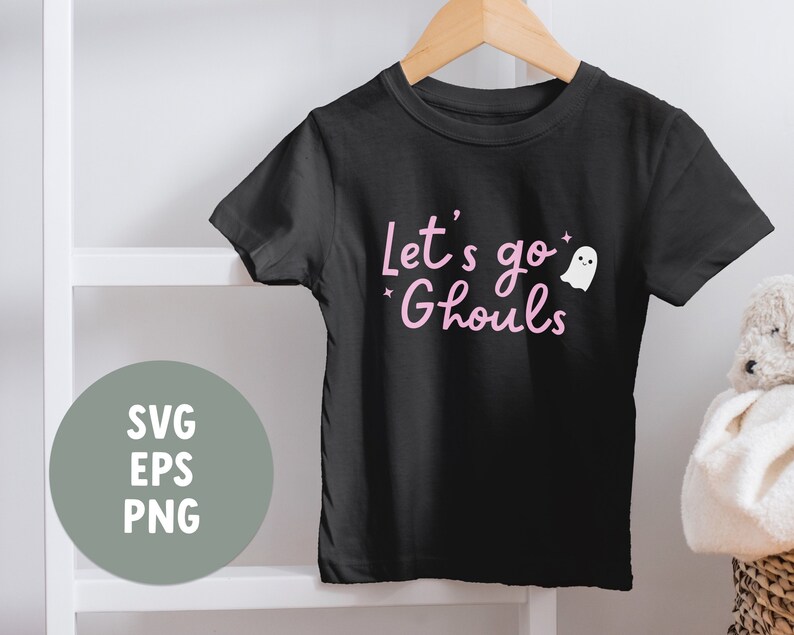 May include: A black short-sleeved t-shirt featuring the pink text Let's go Ghouls and a small white ghost graphic with two tiny stars. The shirt is displayed on a wooden hanger. A circular gray graphic displays the text SVG, EPS, and PNG.