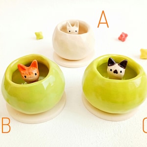 May include: Three ceramic cat figurines in small, round pots. One cat is white, one is orange, and one is black and white. The pots are cream and lime green, with small star-shaped decorations scattered around.