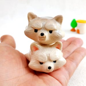 May include: Two small, stacked raccoon figurines. The top raccoon is a light tan colour, and the bottom raccoon is white. Both have black eyes and noses, with a slightly grumpy expression. The figurines are held in a hand.