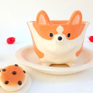 Puede incluir: Una maceta de cerámica con forma de perro corgi, con colores naranja y blanco. La maceta se encuentra sobre un platillo a juego, con dos galletas decorativas cerca. Decoraciones rojas en forma de bayas están dispersas.
