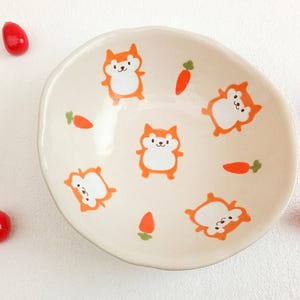 Puede incluir: Un cuenco de cerámica color crema con forma irregular, decorado con zorros y zanahorias de dibujos animados de color naranja. Varios objetos rojos en forma de lágrima están esparcidos alrededor del cuenco. El cuenco es probablemente para servir comida.