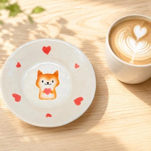 May include: A round, off-white ceramic saucer featuring a hand-painted orange fox character holding a small red heart in its center, surrounded by six scattered red heart motifs of varying sizes, positioned next to a white ceramic coffee mug.