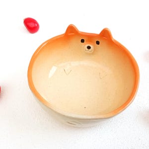 Puede incluir: Un cuenco de cerámica con forma de perro Shiba Inu. El cuenco tiene un interior beige claro y un borde y orejas naranjas. La cara del perro está detallada con ojos y nariz negros. Hay bayas decorativas rojas esparcidas alrededor del cuenco.