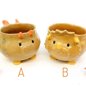 Può includere: Due fioriere a forma di dinosauro in ceramica, una con corna arancioni e macchie verdi, l'altra con corna gialle. Entrambe hanno occhi neri e sono etichettate "A" e "B". Le fioriere sono color cuoio con finitura lucida.