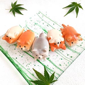May include: Five ceramic animal figurines on a green and white glass plate. The figurines are of a cat, a dog, and three corgis. The corgis are in various poses, including lying down and sitting up. The figurines are all painted in realistic colours.