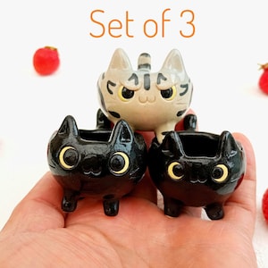 Puede incluir: Juego de tres macetas de cerámica con forma de gato. Dos macetas de gato negro y una maceta de gato gris y blanco. Las macetas tienen ojos amarillos y están rodeadas de fresas rojas.