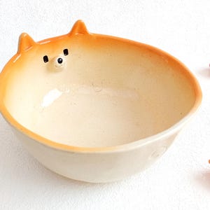 Puede incluir: Un cuenco de cerámica con forma de perro Shiba Inu. El cuenco es de color crema con detalles en naranja y presenta las orejas y la cara del perro. Objetos decorativos rojos están dispersos alrededor del cuenco.