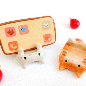 Puede incluir: Un caprichoso soporte de cerámica con forma de teléfono con iconos de aplicaciones con temática de gatos, un soporte blanco con forma de conejito y una figura de gato atigrado naranja. Objetos rojos en forma de lágrima están esparcidos.