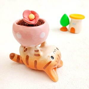May include: A ceramic orange and white cat figurine holding a pink planter with a pink flower on top. The planter has white polka dots.