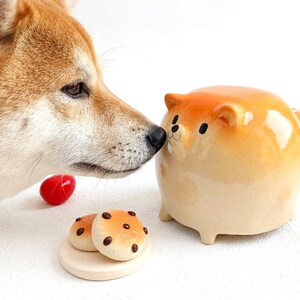 Puede incluir: Un perro Shiba Inu olfatea un recipiente de cerámica con forma de perro con un degradado naranja y crema. Dos pequeños objetos redondos de color crema con manchas marrones se encuentran sobre una base de madera, y objetos rojos en forma de corazón están dispersos.