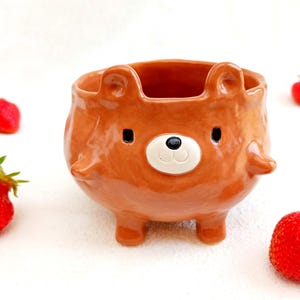 Puede incluir: Una maceta de cerámica marrón con forma de oso con cara blanca y negra, orejas pequeñas y patas cortas. Fresas rojas están esparcidas alrededor de la maceta sobre una superficie blanca. La maceta está diseñada para contener plantas pequeñas o suculentas.