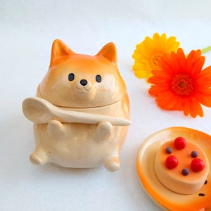 Könnte beinhalten: Eine orangefarbene und weiße Keramik-Zuckerdose in Form eines Hundes mit einem Holzlöffel. Die Schüssel hat schwarze Augen und eine braune Nase. Die Schüssel steht auf einer weißen Oberfläche.