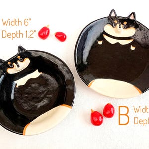 Puede incluir: Dos cuencos de cerámica con forma de Shiba Inu. El cuenco A mide 15 cm de ancho y 3 cm de profundidad. El cuenco B mide 17 cm de ancho y 1,5 cm de profundidad. Ambos cuencos son negros con detalles beige y blancos. Dos decoraciones rojas en forma de baya están junto a cada cuenco.