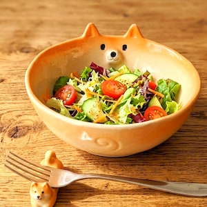 May include: A ceramic bowl shaped like a Shiba Inu dog, featuring a light orange exterior, pointed ears, and a small face on the rim, filled with a fresh salad of lettuce, tomatoes, and cucumbers, accompanied by a metal fork resting on a dog-shaped holder.