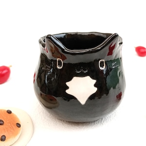 May include: A glossy black ceramic cat-shaped container with a white emblem on the front. The container has two silver accents and a wide opening. Red decorative berries and a cookie are scattered around the container.