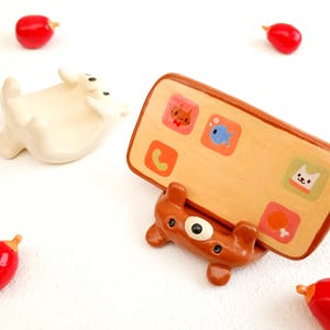 May include: A brown ceramic phone stand shaped like a bear, holding a tan phone with colorful app icons. A white ceramic animal figure and several red, teardrop-shaped objects are scattered around.