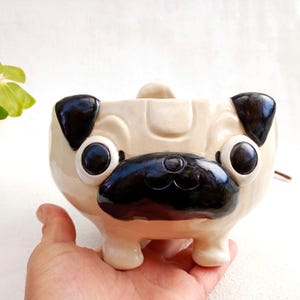 May include: A ceramic planter shaped like a pug dog with black ears, nose, and eyes. The planter is beige with a glossy finish.