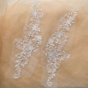 May include: Two white lace appliques with floral designs and silver sequins on a beige background.