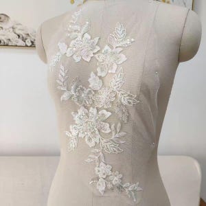 Könnte beinhalten: Weiße florale Spitzenapplikation mit Pailletten und Perlen. Die Applikation zeigt ein Design aus Blumen und Blättern und wird auf einer Schaufensterpuppe präsentiert. Die filigranen Details und schimmernden Akzente erzeugen einen romantischen und eleganten Look, passend für Braut- oder Abendmode.