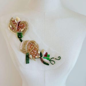 May include: A floral-shaped applique patch featuring intricate beadwork in shades of gold, pink, and green. The design includes sparkling clear crystals, metallic gold seed beads, and vibrant green glass beads arranged in a botanical pattern.
